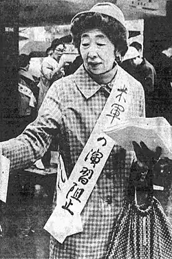 Portrait of an Asian woman in a hat wearing a checked coat draped with a sash bearing a Japanese slogan on her left shoulder and carrying a large bag in her left hand.