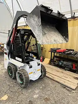 Yuchai S35 electric skid steer loader with raised bucket, photographed at a heavy equipment yard. The compact, battery-powered machine is positioned on a gravel surface near tools and a workbench.
