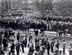 Several police officers on horses watch a large crowd standing in a public park.