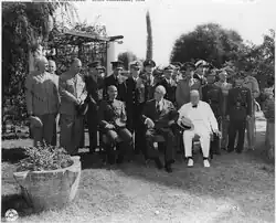 Black and white photograph. Three men are seated on chairs in a garden, the one at the right in a white suit, bowtie and a hat placed on his right leg; the one at the centre is in a suit and tie; the one at the left is in army uniform with a cap placed on his left leg. Multiple men in varying military uniforms stand behind them